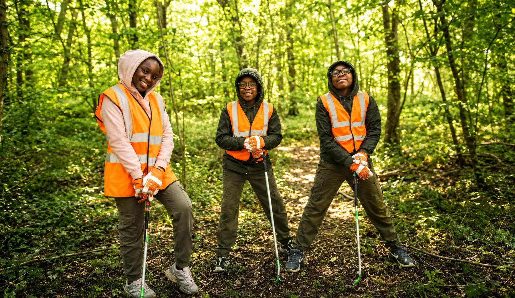 Planet Trio — Waimi, Mbetmi and Yimi in high-visibility vests with litter pickers in a sunlit forest